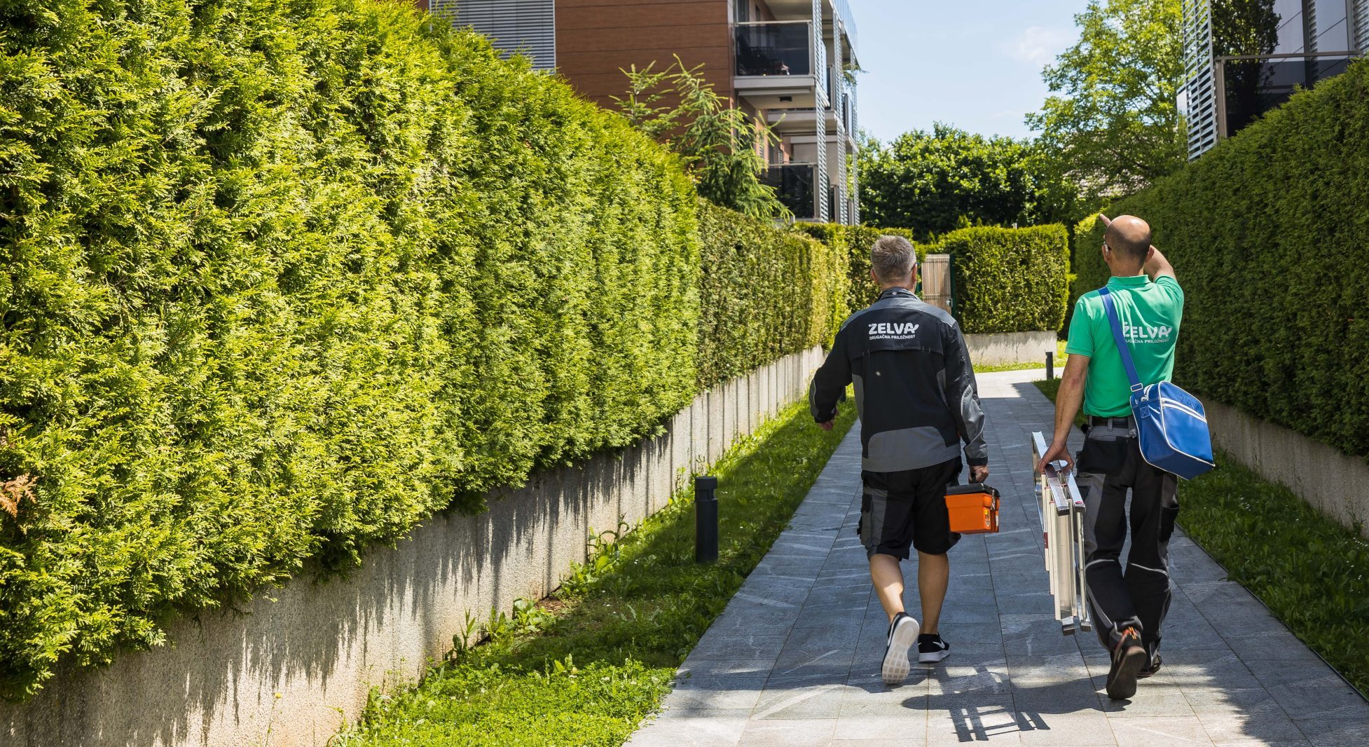 Dva delavca v uniformah na poti prosti stanovanjski stavbi predstavljata Tehnične storitve.
