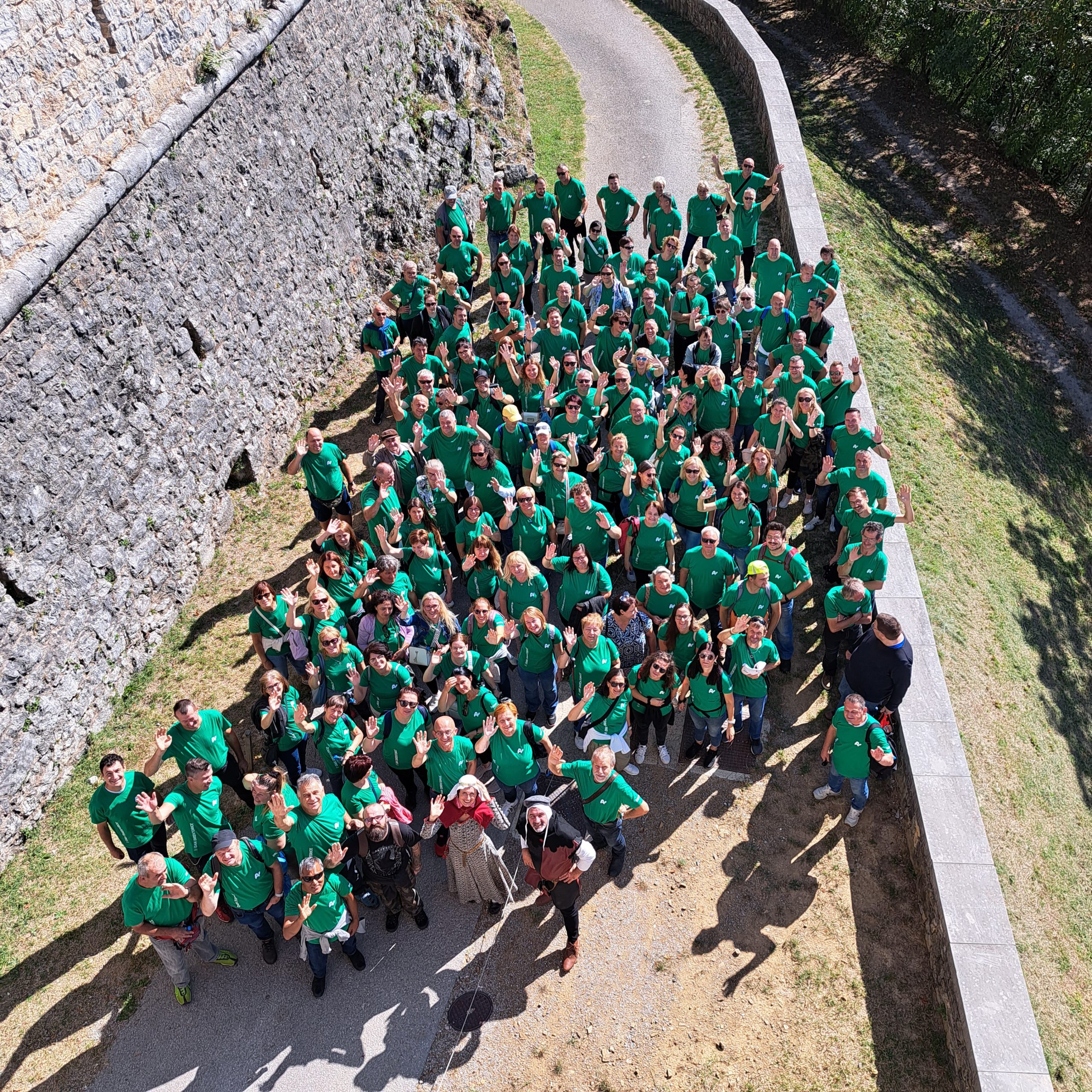 O nas - skupinska fotografija velike skupine Želva zaposlenih v zelenih majicah, ki pozira pred kamnitim zidom.