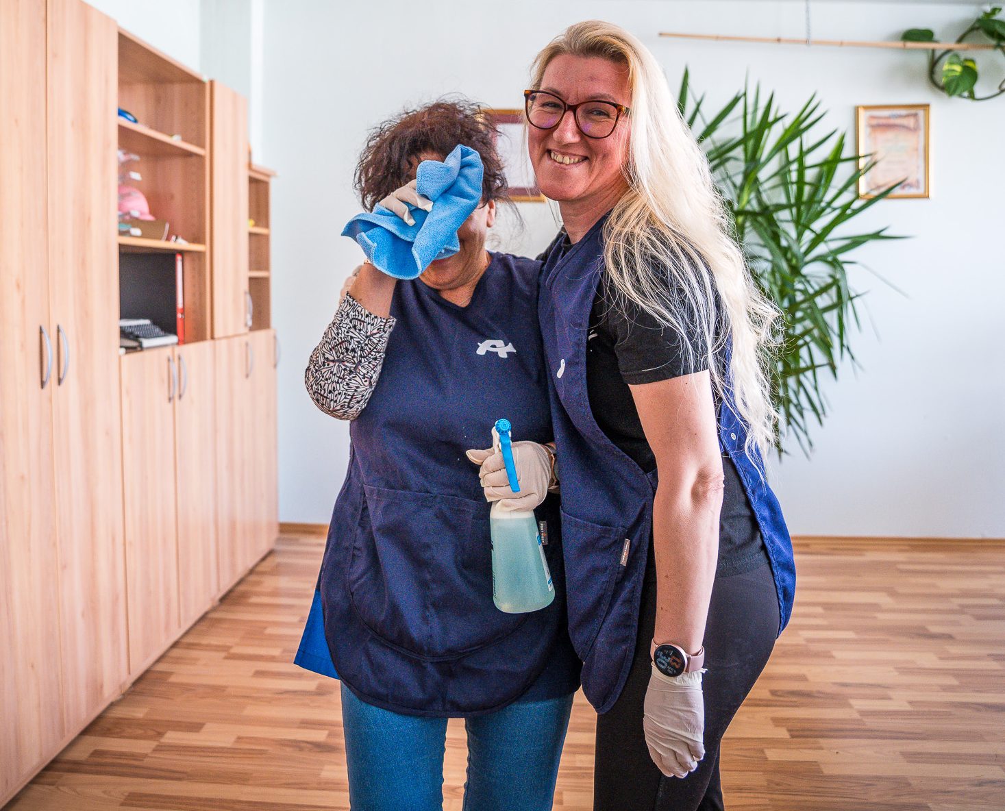Dve nasmejani sodelavki v modrih uniformah čistilnega servisa izvajata čiščenje pisarne.
